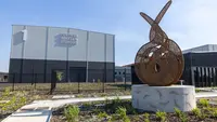 A photo of Perth Film Studios with a large bronze abstract sculpture in the landscaped forecourt, modern studio buildings, perimeter fencing, and a clear blue sky.