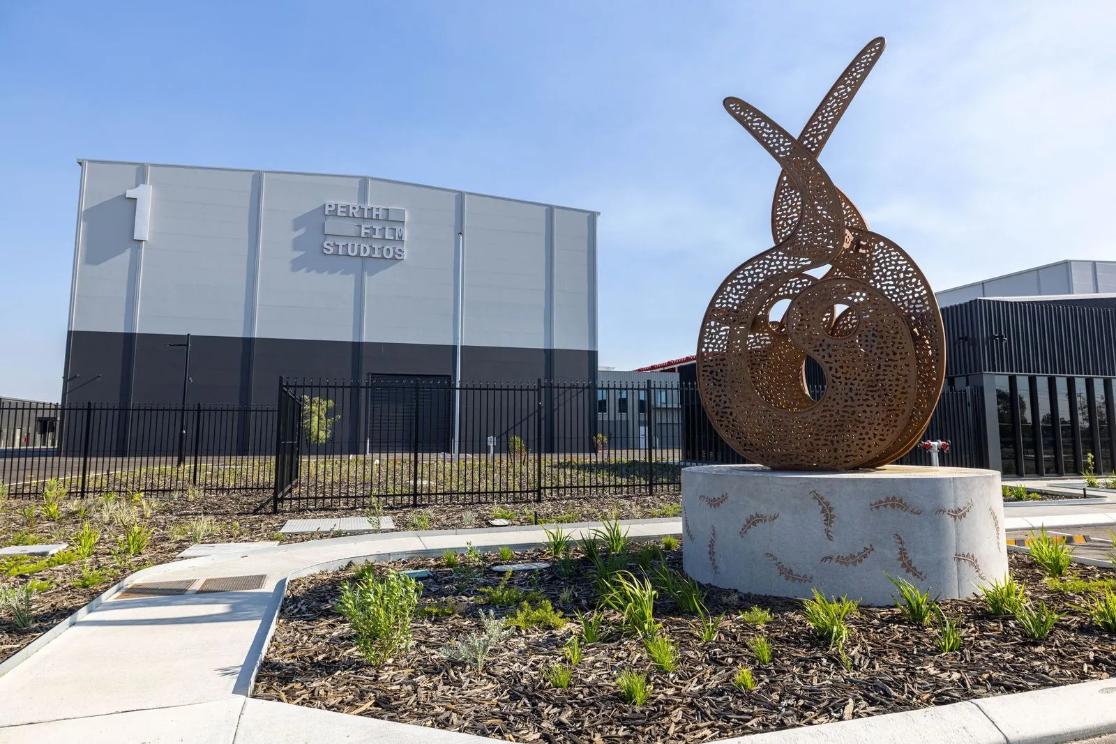 A photo of Perth Film Studios with a large bronze abstract sculpture in the landscaped forecourt, modern studio buildings, perimeter fencing, and a clear blue sky.